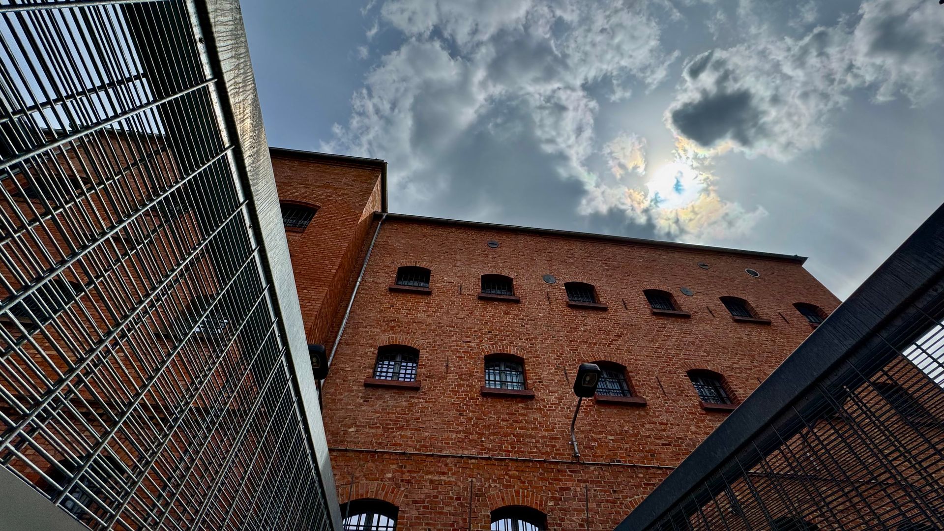 View upwards between metal grilles towards the red-brick prison building, barred windows and a cloudy sky at Andreasstraße in Erfurt.